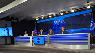 people in suits sit at a desk in front of a poster of four astronauts in flight suits and the text "crew-8"
